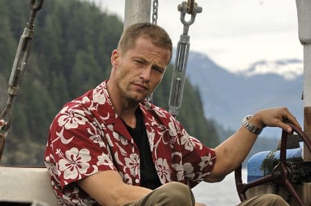 A man with short hair wearing a red Hawaiian shirt sits at the helm of a small boat, looking intently ahead. The backdrop features lush green hills and a cloudy sky, creating a serene but rugged coastal scene.