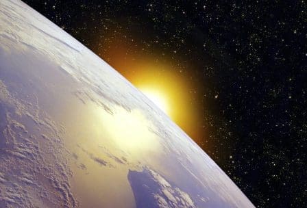 A view of Earth from space, capturing the curvature of the planet and the golden glow of the sun rising above the horizon, set against a backdrop of twinkling stars in the dark cosmos.