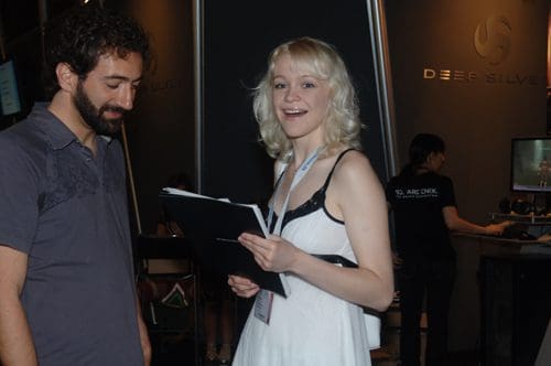 A smiling woman in a white dress holds a clipboard, speaking to a man in a gray shirt at an event booth labeled 'Deep Silver'. The background features other attendees and screens displaying content.