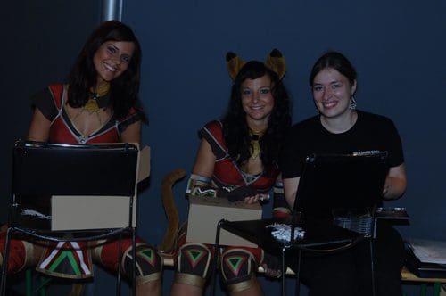 Three women, two in vibrant costumes resembling ancient warriors with animal ears, sit side by side, smiling at the camera. The third woman, dressed casually, is seated with a laptop. They are indoors, against a dark blue backdrop.