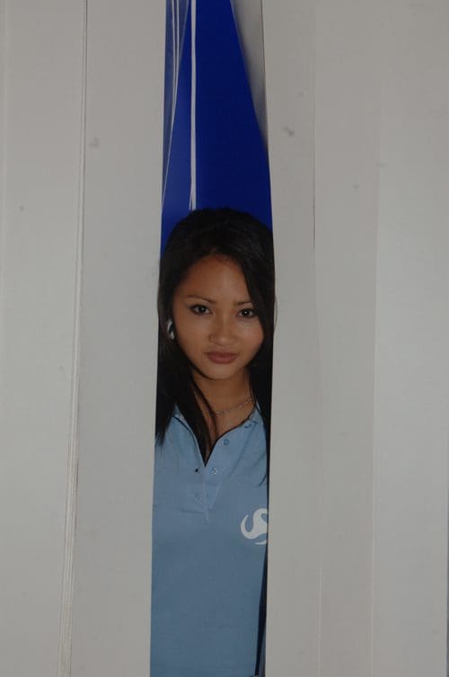 A young woman peeks through a narrow gap between two white panels, wearing a light blue polo shirt. The background features a vibrant blue, creating a striking contrast with her dark hair and expression.