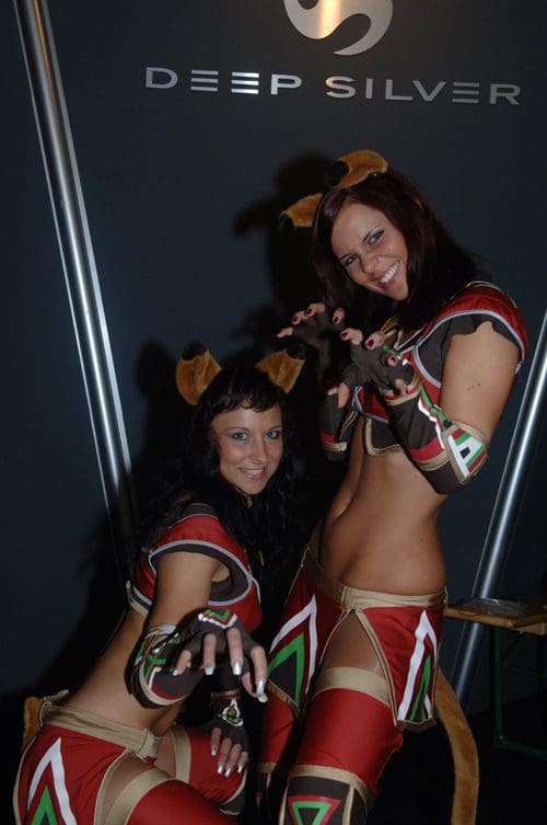Two women in tribal-inspired costumes with animal ears posing playfully at an event. They are wearing colorful outfits that feature patterns and cutouts, with a background displaying the Deep Silver logo.
