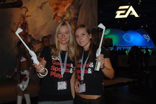 Two women smiling, wearing black T-shirts with a gaming slogan, holding white gaming controllers, in a lively event space with game banners and colorful lighting in the background.