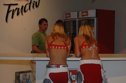 Two women in matching red and white outfits with bare backs stand at a counter, engaging with a man in a green shirt behind the bar. The background displays a logo for 'Fructiv,' and there's a beverage cooler visible.
