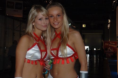 Two female models wearing matching silver and red outfits with fringe details, smiling and posing for a photo in a well-lit indoor setting, with blurred displays in the background.
