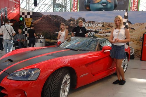 A striking red sports car with black racing stripes displayed at an exhibition. Two women in white tops and skirts stand beside the car, holding checkered flags. A backdrop features a desert landscape and promotional signage.