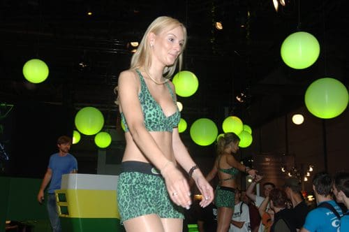A model walking on a stage wearing a green leopard print bikini and shorts. The background features large green spheres suspended from the ceiling, with spectators watching the fashion event.