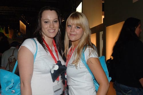 Two young women pose for a photo at an event, both wearing matching white t-shirts with event badges. They have long hair and are smiling, standing close together with colorful backpacks.