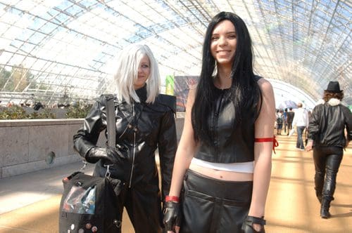 Two women in cosplay posing together in a large glass atrium. One has short white hair and wears a black leather jacket; the other has long black hair, wears a white top, and a black leather skirt. People and plants are visible in the background.
