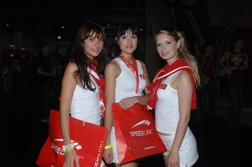 Three women posing together in matching outfits, each holding a red promotional bag with 'SPEEDLINK' printed on it. They are indoors at a crowded event, with a casual, friendly atmosphere.