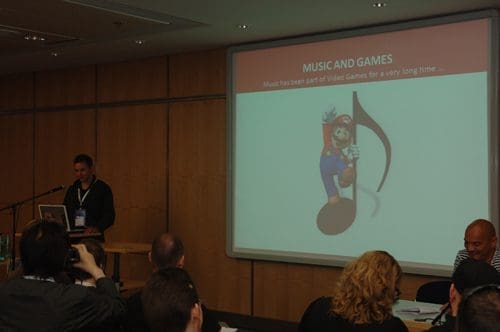 A speaker presenting at a conference with a projector displaying the title "MUSIC AND GAMES". An animated character resembling a plumber stands beside a large musical note on the screen. Attendees are seated in the foreground.