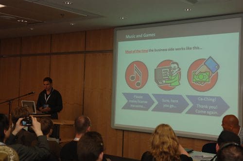 A speaker presenting at a conference, standing at a podium with a laptop. Behind him, a slide shows three graphics related to music and games. An audience of engaged attendees is visible in the foreground, listening attentively.