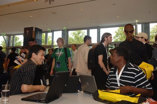 A diverse group of people congregating in a spacious event venue, with some individuals engaged in conversations, while others sit at a table working on laptops. The atmosphere suggests a tech conference or workshop.