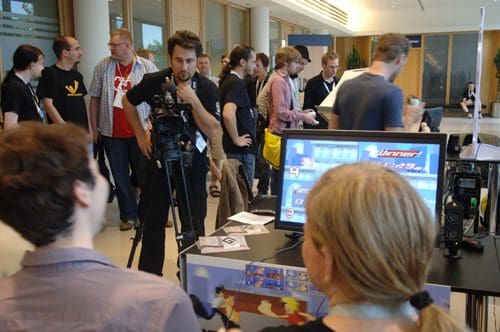 A group of people gathered at an event, with two individuals in the foreground examining a gaming setup. One person is using a camera, while the others are engaged in conversation, showcasing a vibrant atmosphere.