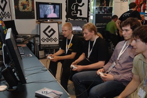 Four young men sitting in front of computer monitors, playing video games at a gaming convention. Each is holding a controller, focused on the screens. In the background, a logo is visible on a wall.