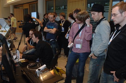 A group of people gathered around a computer station at an event, engaging in discussions. Some are observing the screen, while others appear to be interacting with equipment, showcasing interest and collaboration.