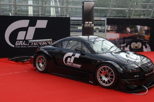 A sleek black racing car displayed on a red carpet, featuring the Gran Turismo logo on its side. The background includes banners and screens showcasing racing visuals.