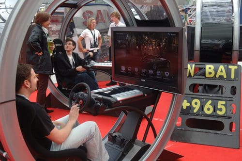 A group of people observing an interactive racing simulator at an exhibition. A man is seated in a simulator with a steering wheel, while a large screen displays a racing game. A vibrant red carpet adds color to the setting.
