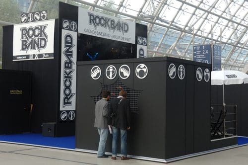 A promotional booth for the game "Rock Band" featuring bold logos and musical icons. Two men are interacting with the display, set in a spacious, bright indoor area with a modern architecture background.