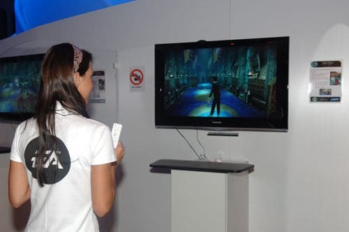 A young woman with long hair wearing a white t-shirt stands in front of a television screen displaying a video game. The screen shows a character navigating through a vibrant and colorful game environment.