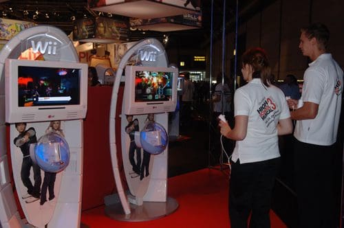 Two people playing interactive video games on Wii consoles at an event. The setup features large screens displaying colorful graphics, surrounded by bright red flooring and vibrant event decorations.