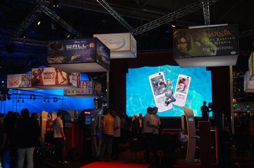 A bustling video game exhibition with colorful booths displaying titles like Disney's "Wall-E" and "High School Musical." People are interacting with gaming stations while large screens showcase game visuals.