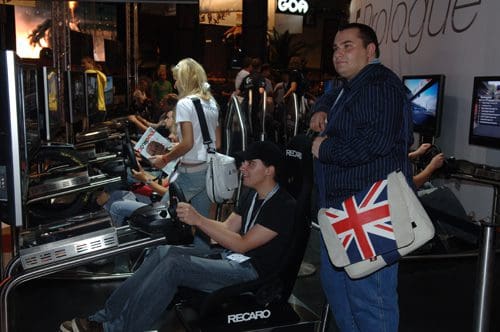 A gaming event scene featuring multiple people engaged in racing simulation games. One player, focused on the screen, is seated in a racing chair, while another man stands nearby, wearing a blue striped shirt and carrying a bag with a British flag.