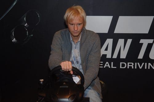 A man with short blond hair focused on driving a racing simulator, holding a steering wheel. He wears a gray cardigan and denim jeans, sitting against a dark background with graphics suggesting a racing theme.