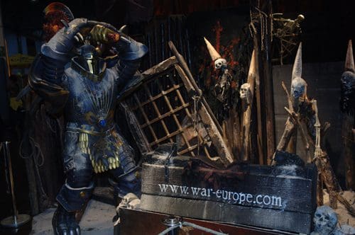 A life-size armored knight wielding a sword stands in a spooky display, surrounded by eerie skeletal figures and a dilapidated wooden structure. The scene is dark and atmospheric, enhancing the theme of a haunted setting.