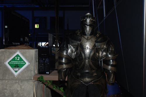 A full suit of medieval armor displayed beside a concrete block with a green safety sign. The armor features intricate designs and a polished finish, set against a dark background with faint lighting.