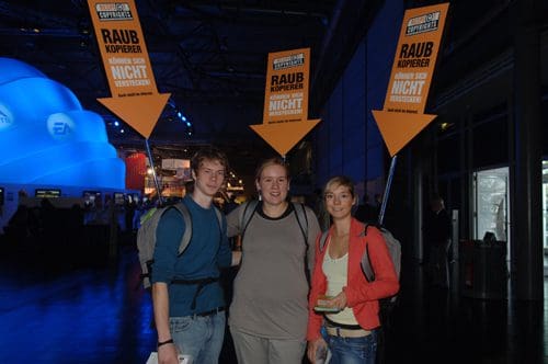 Three young adults standing indoors, holding signs that read "RAUB KOPIERER" with arrows pointing down. The background features an eye-catching blue structure and illuminated areas, suggesting an event atmosphere.