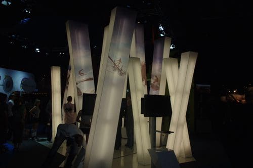 An artistic installation of tall, illuminated pillars arranged in a cluster, featuring images printed on their surface. People walk around the installation in a dimly lit exhibition space with blue ambient lighting.