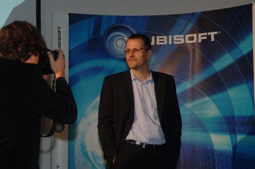 A man in a black suit and light blue shirt poses while a photographer captures his image at a promotional event, with a blue and white Ubisoft backdrop featuring swirling graphic designs.
