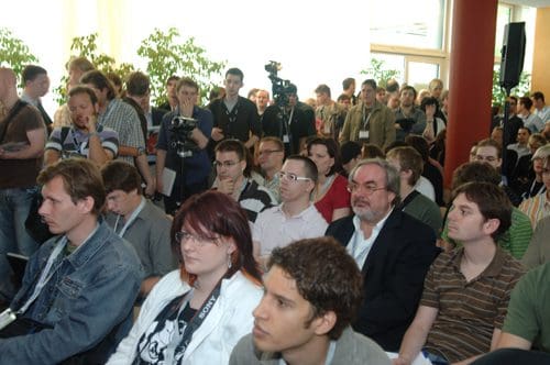 A crowd of attendees seated at a conference, with individuals engaged in discussion or listening. Some people are looking at the stage, while others are focused on their devices. The room is bright and filled with greenery.