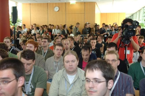 A large audience of attendees seated in a conference hall, focused on a presentation. Photographers are capturing the moment, while a clock is visible on the wall. Most attendees wear lanyards and exhibit various expressions of concentration.