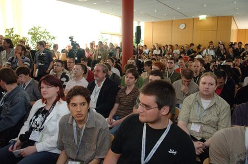 A large audience gathered in a conference hall, seated row by row. Attendees of diverse backgrounds, including individuals with cameras, watch intently towards the front, where a presentation or event is taking place.