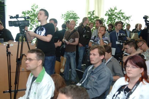 A diverse group of people attending a conference, some taking notes and others recording with cameras. Green plants are visible in the background, creating a lively atmosphere.