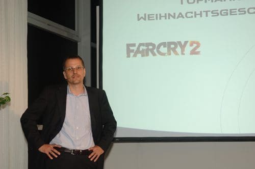 A man wearing glasses and a black suit with a light blue shirt stands confidently with his hands on his hips. Behind him is a presentation screen displaying the title 'Far Cry 2'.