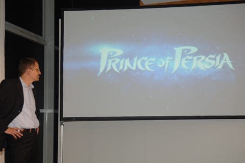 A presenter stands beside a large screen displaying the title "Prince of Persia" in stylized lettering, set against a starry background during a presentation.