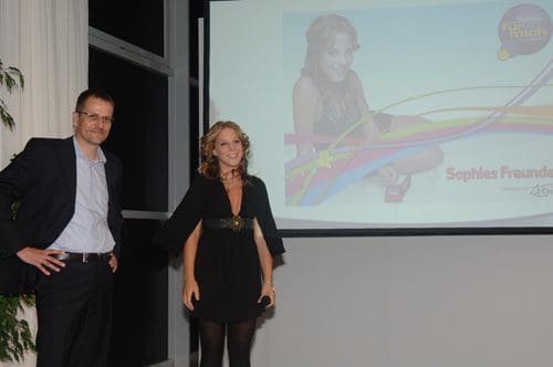 Two presenters standing in front of a large screen during an event. The man, wearing glasses and a dark suit, poses next to a woman in a black dress with flowing sleeves. The screen features a colorful design and the name 'Sophies Freundt'.