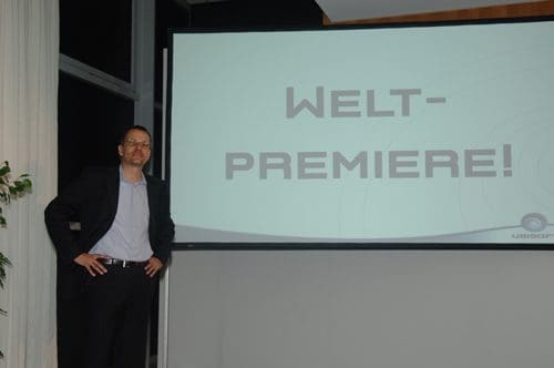 A man in a black suit and white shirt stands next to a large screen displaying the words "WELT-PREMIERE!" in bold letters during an event, with a modern interior design in the background.