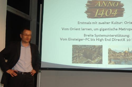 A presenter standing beside a screen displaying the game title 'Anno 1404' and text in German discussing gameplay, including cultural aspects and system requirements. The presenter appears engaged, wearing a dark blazer and light shirt.