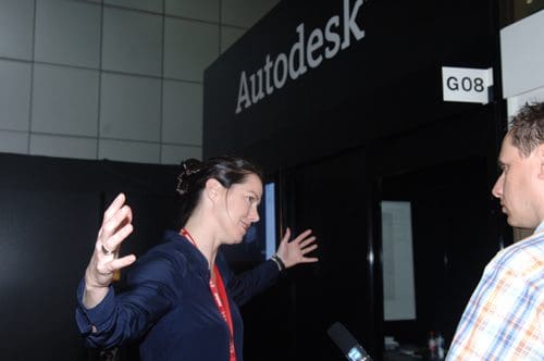 A woman with dark hair gesturing expressively while speaking to a man near an Autodesk booth at an event. The backdrop features the Autodesk logo and a booth number, with a modern interior setting.