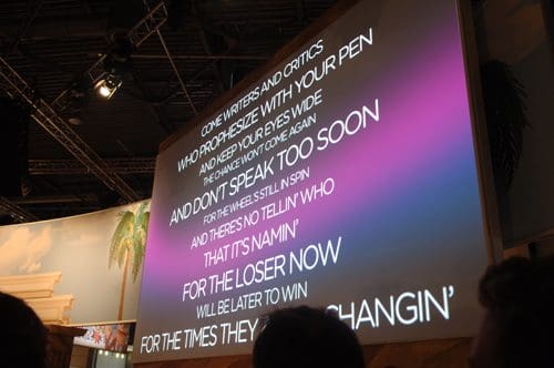 A large projection screen displaying lyrics with vibrant colors in a dimly lit venue. The text encourages writers and critics to open their eyes, highlighting themes of change and resilience.