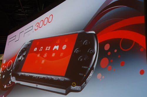 A promotional display featuring the PSP 3000 portable gaming console, with a vibrant red background. The console is shown with a bright screen displaying icons for media and games.