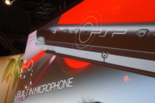 Close-up of a gaming console with a sleek design, showcasing buttons and a built-in microphone feature. The background features an eye-catching gradient with circular patterns and text indicating 'BUILT-IN MICROPHONE'.