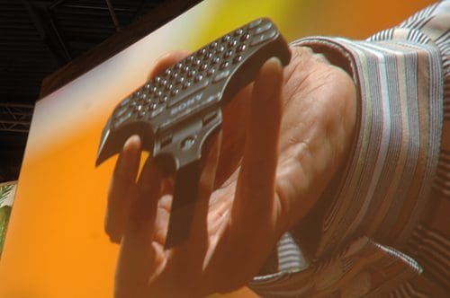 A person's hand holding a small, black electronic device with a textured surface, displayed against a vibrant orange background.