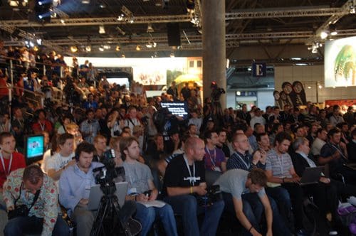 A large crowd of attendees seated in an auditorium during an event. Various individuals are focused on a presenter, with cameras and laptops set up in front. Bright lights and screens illuminate the space, reflecting a lively atmosphere.