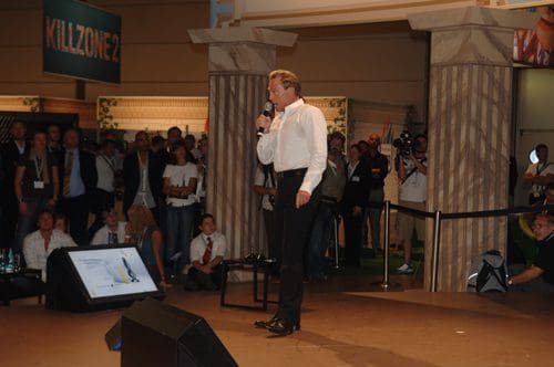 A speaker in a white dress shirt stands on a stage, holding a microphone and addressing an audience. In the background, a crowd watches attentively, with banners visible, including one that reads 'KILLZONE 2.' The setting appears to be an event or convention.