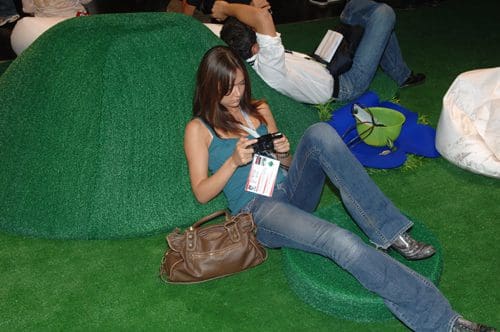 A woman wearing a blue tank top and jeans sits on a green, cushioned surface, focused on using a camera. In the background, another person relaxes on a larger green mound, with various bags and items scattered around on the artificial grass floor.